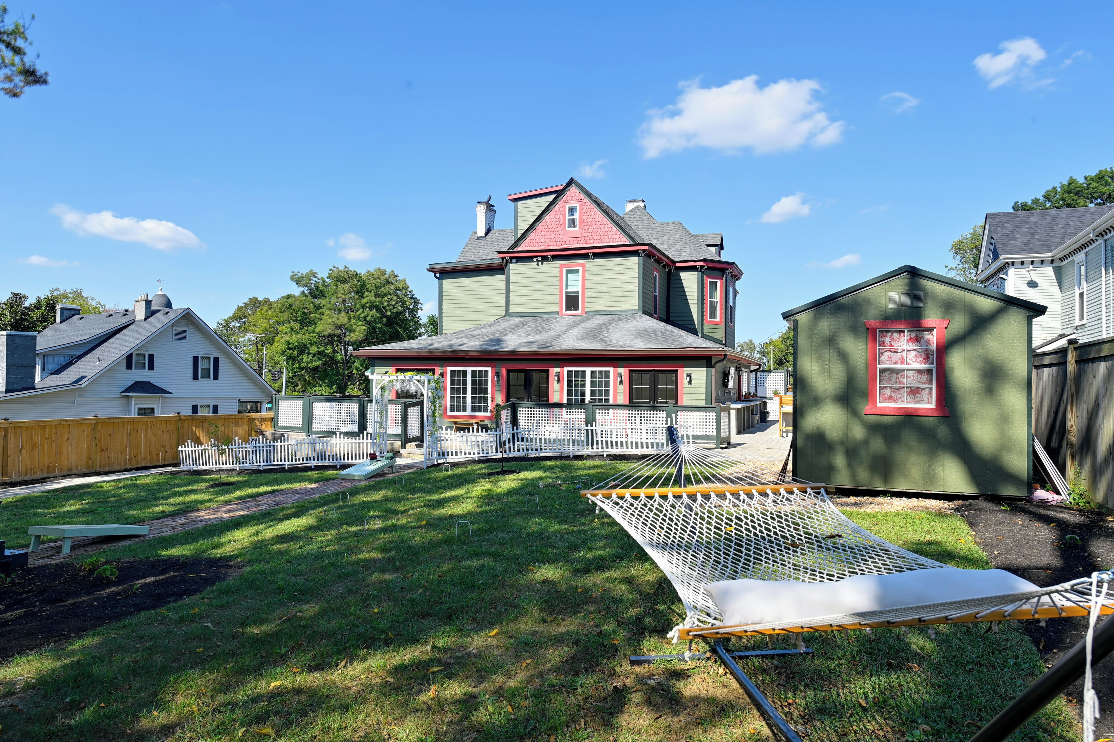 A large house with a red roof and green siding, surrounded by a white picket fence and a hammock.