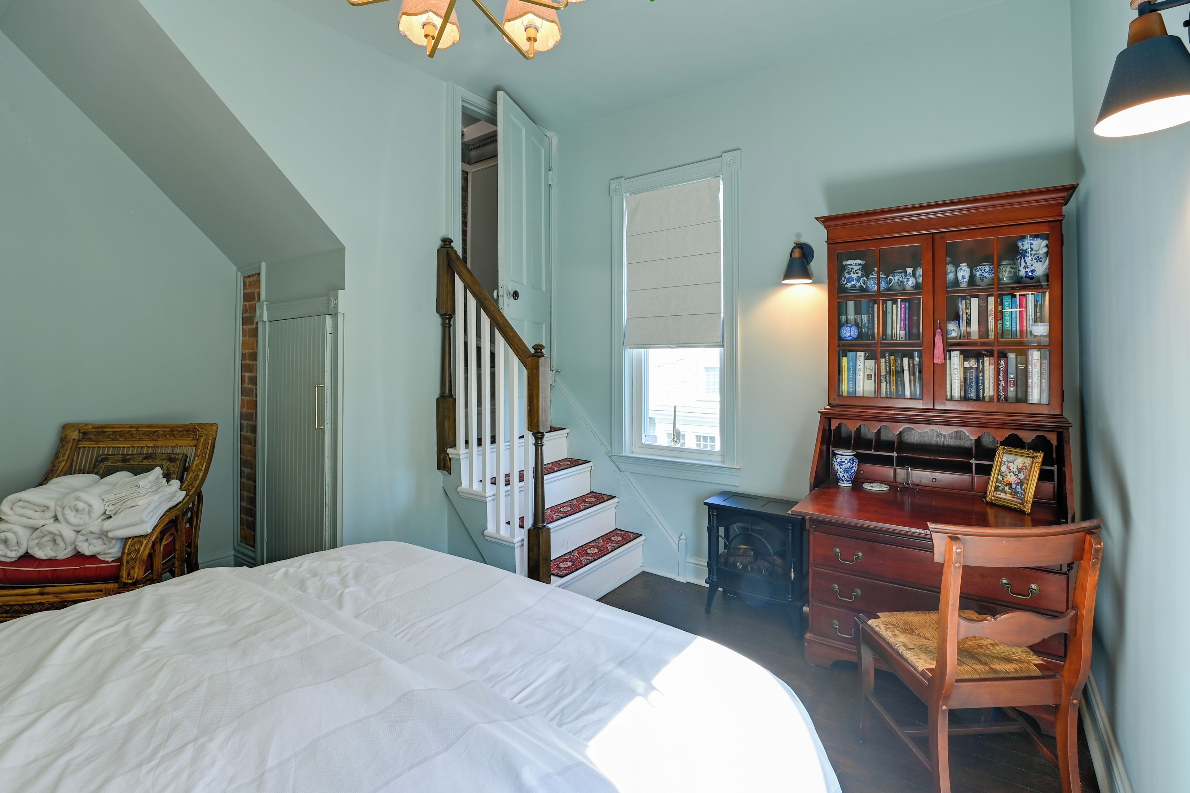 A bedroom with a bed, a wooden chair, a bookshelf, a fireplace, and a staircase.