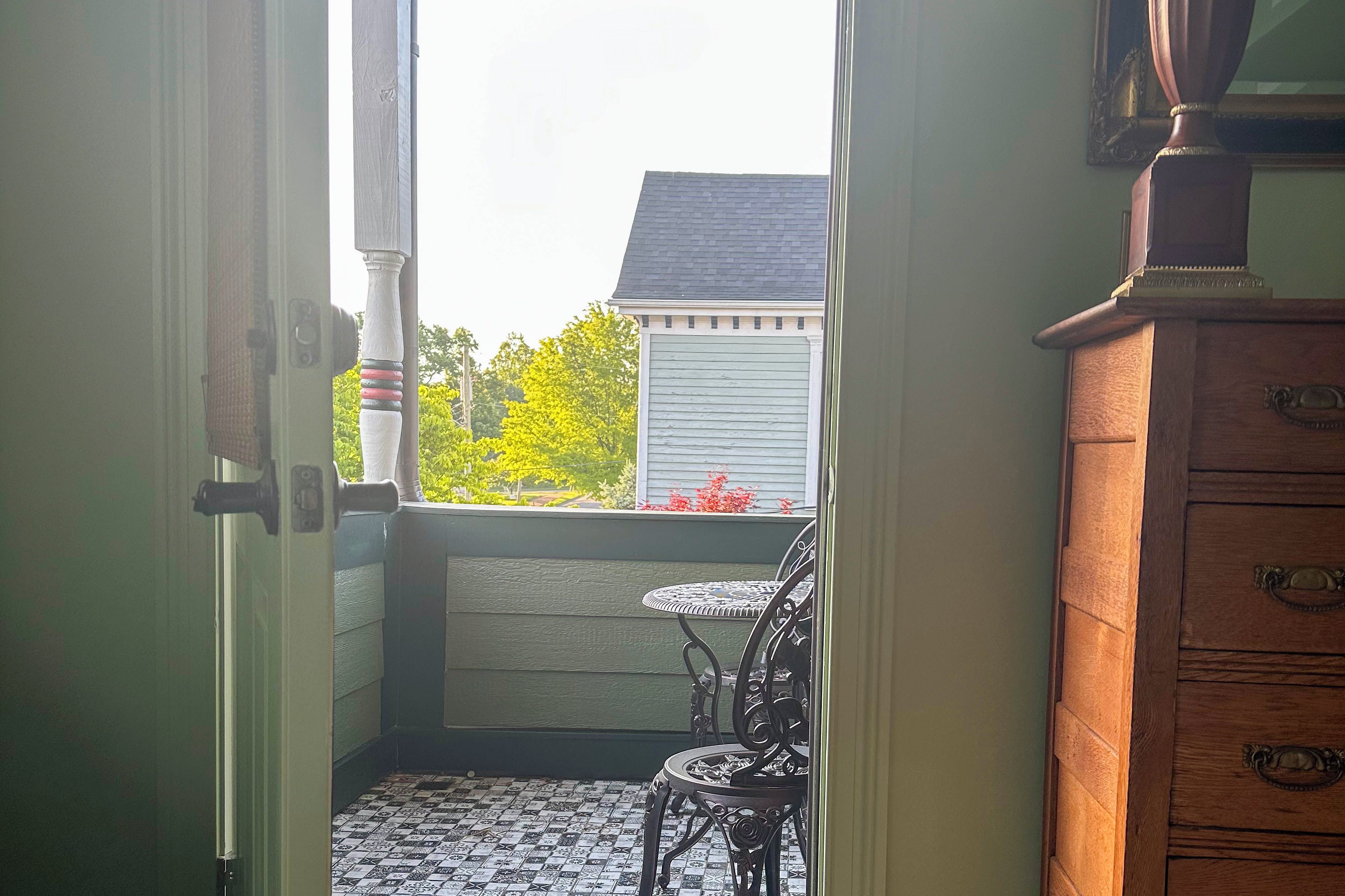 A view from a room's door to a balcony with a table and chairs, a wooden dresser, and a window overlooking a house and trees.