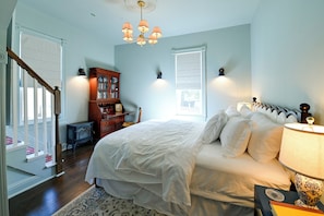 A bedroom with a large bed, a wooden dresser, a chest of drawers, a small table, and a lamp.