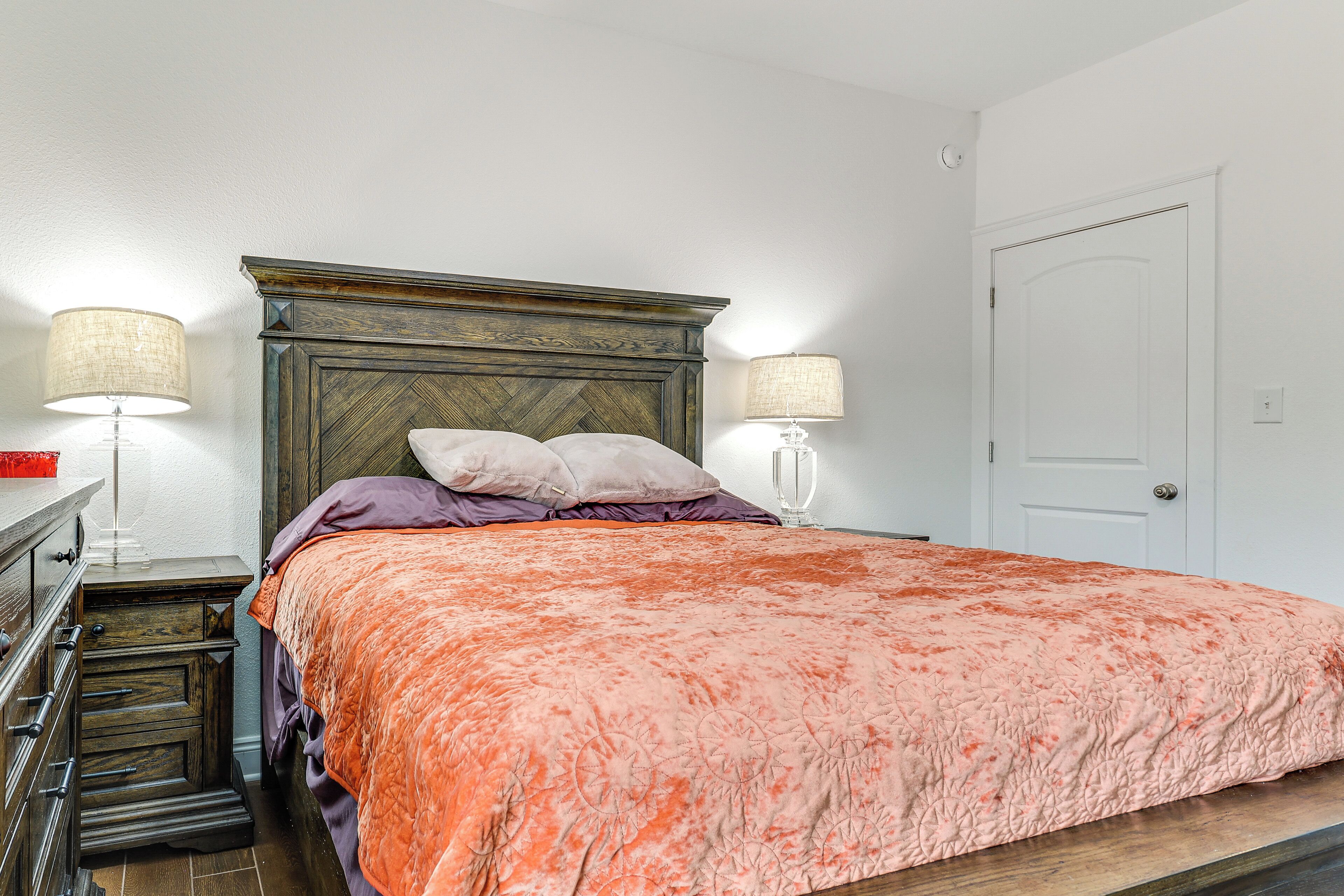 A bedroom with a large bed, two bedside lamps, a wooden dresser, and a door.