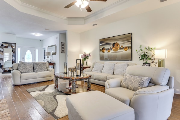 A spacious living room with a high ceiling, a ceiling fan, and a large painting. There are two sofas and a coffee table with decorative items.