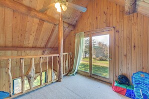 A wooden cabin interior with a ceiling fan, a large window with a view of the outdoors, and a small fan on the floor.