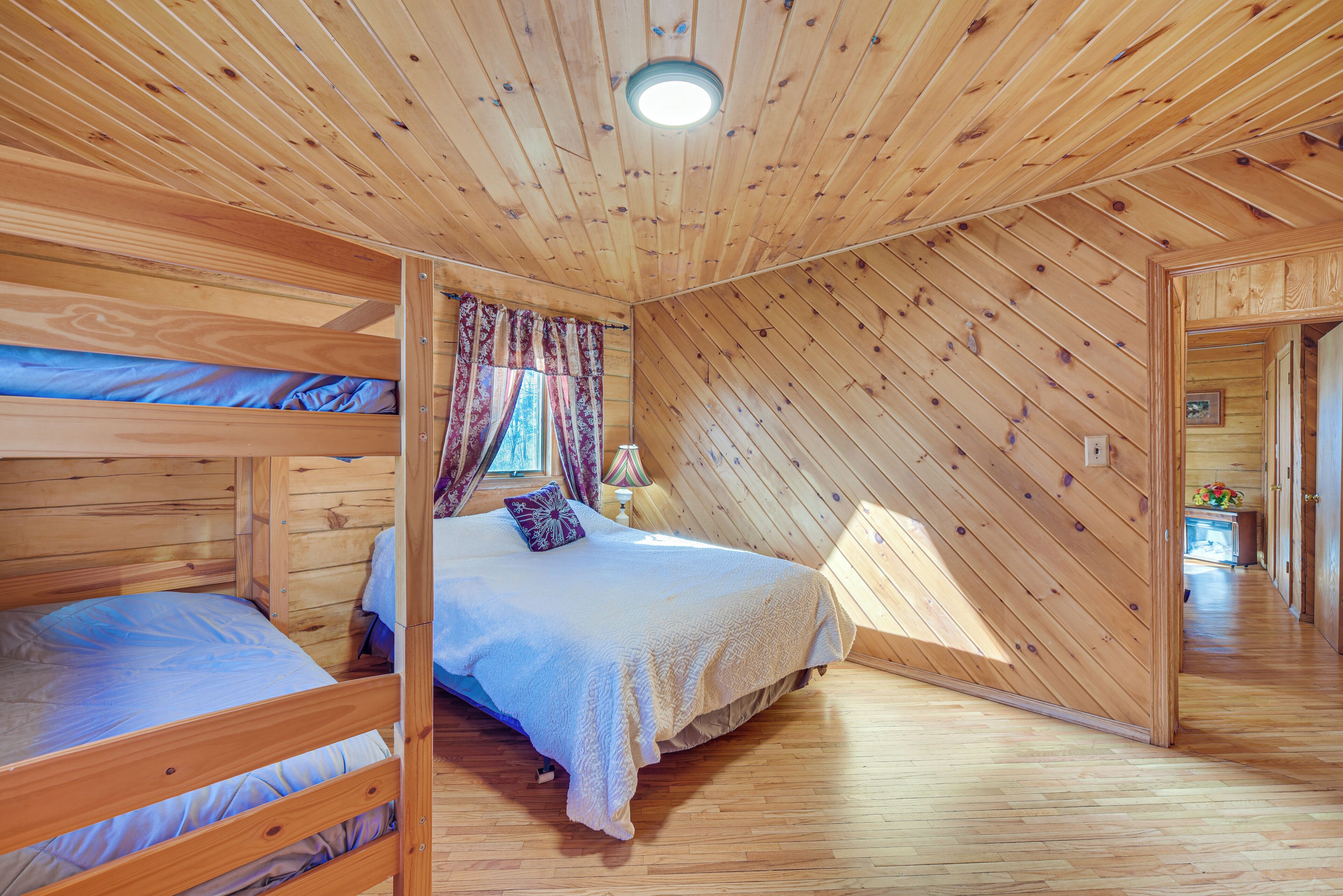 Un dormitorio de cabaña de madera con literas, una cama con una colcha blanca, una mesita de noche y una ventana con cortinas.