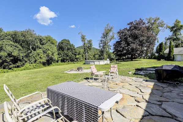 A stone patio with outdoor furniture, a grill, and a view of a lush green landscape with trees.