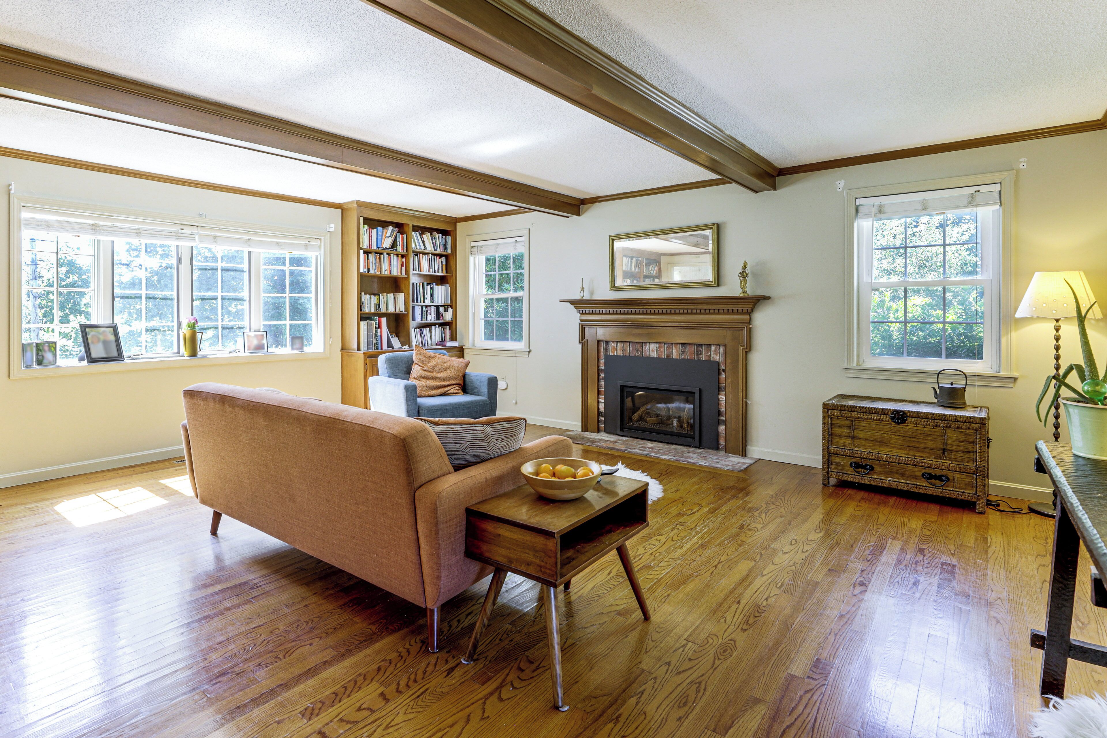 A living room with hardwood flooring, a fireplace, a bookshelf, and a window with a view of trees.