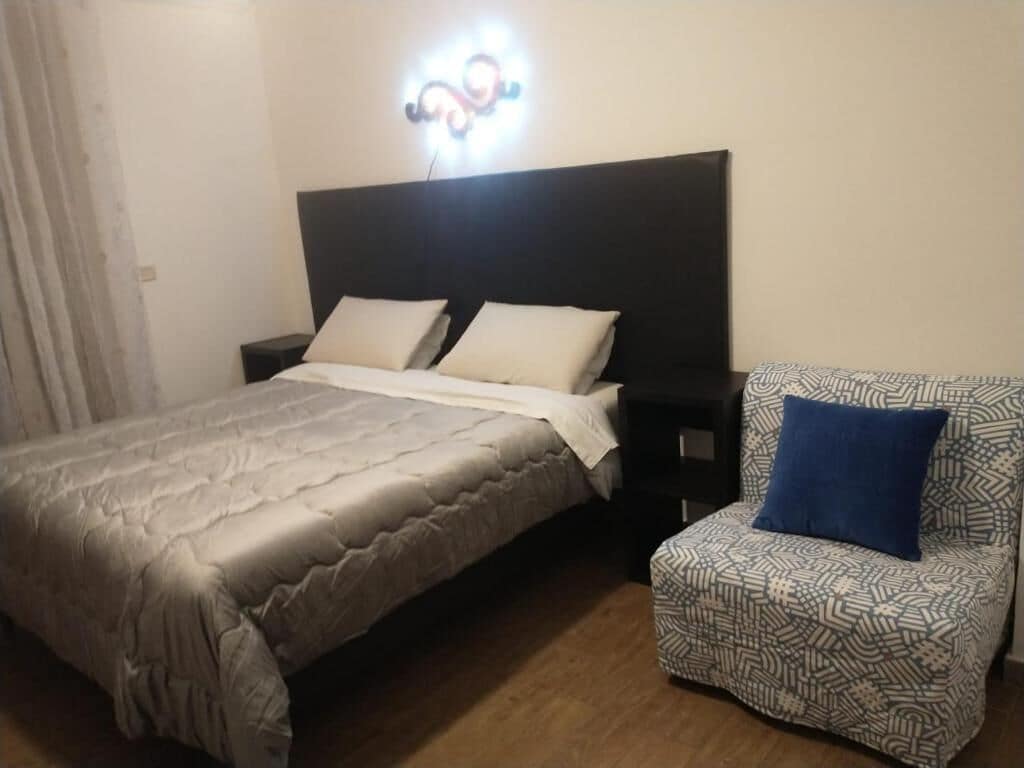 A bedroom with a bed, a nightstand, a patterned armchair, and a wall-mounted light fixture.