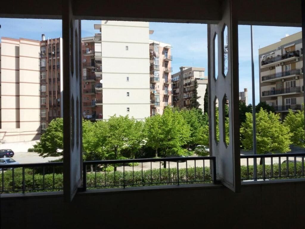 A view from a window with a balcony, overlooking a row of multi-story residential buildings and a green area with trees.