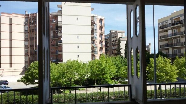 Blick aus einem Fenster mit Balkon auf eine Reihe mehrstöckiger Wohnhäuser und eine Grünfläche mit Bäumen.