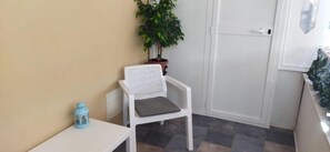 Una habitación pequeña con una silla blanca, una mesita, una planta en maceta y una puerta.