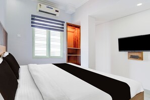 A hotel room with a bed, a TV mounted on the wall, an air conditioning unit, a window with blinds, and a built-in shelf.