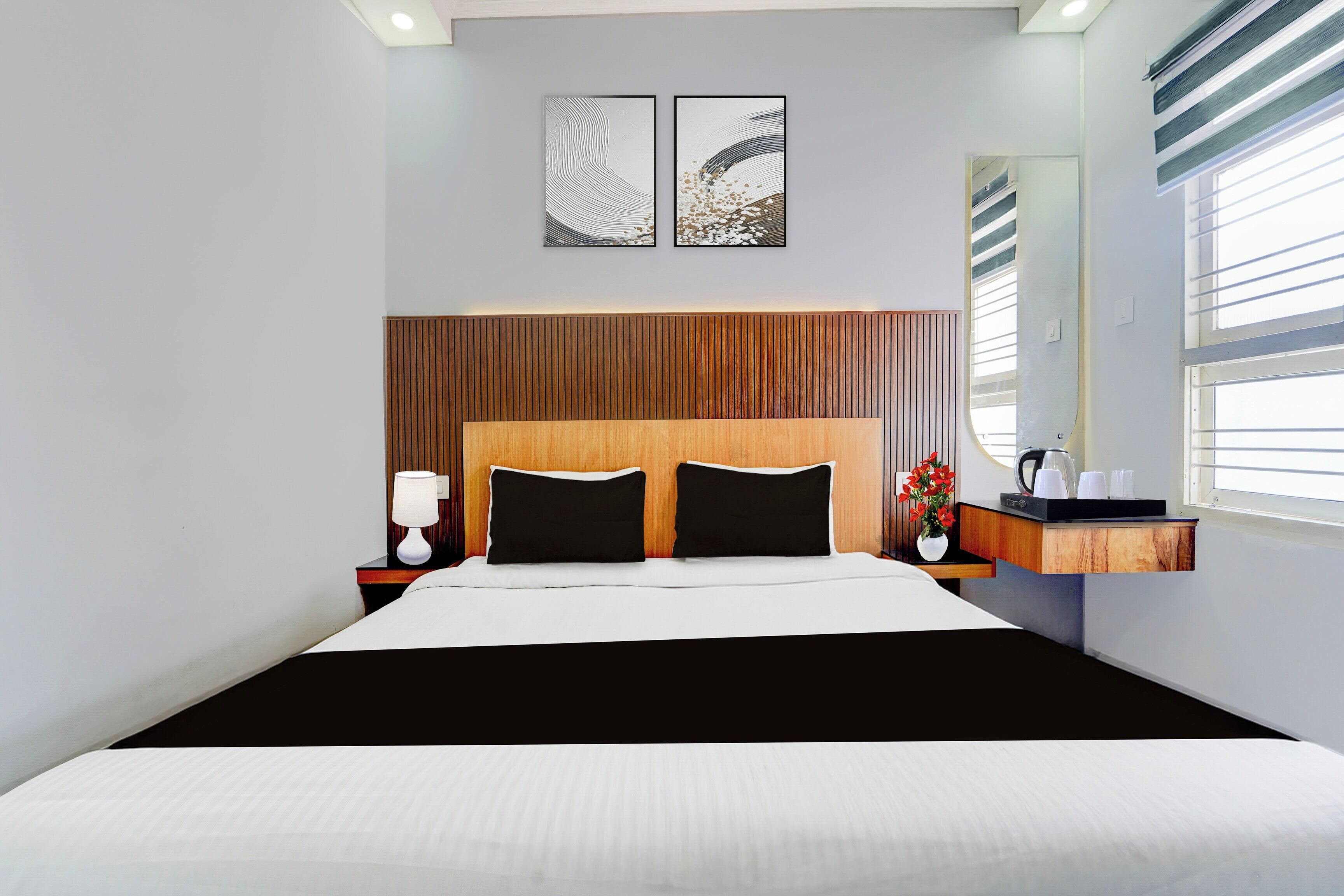 Una habitación de hotel moderna con una cama grande, una mesita de noche con lámpara, una pequeña mesa con un jarrón de flores y una ventana con persianas.