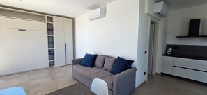 Ein moderner Wohnbereich mit grauem Sofa, weißem Sessel, einer Küchenzeile und einem Kleiderschrank mit viel Stauraum.