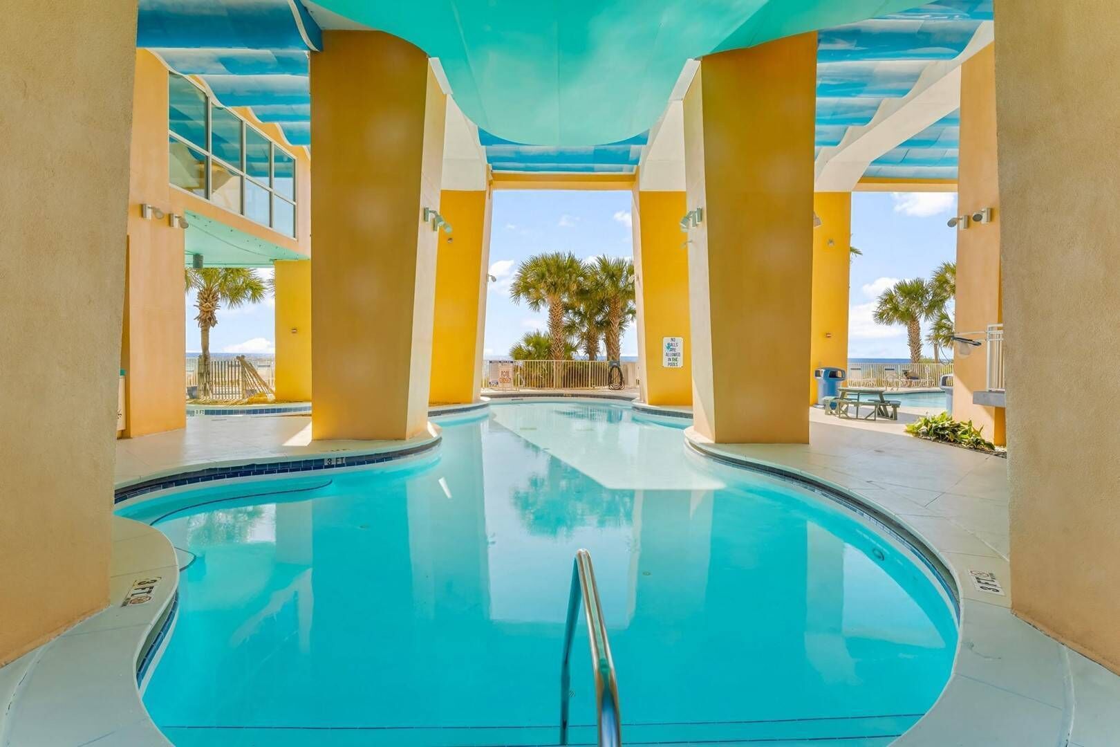Piscina com água azul cristalina, rodeada por paredes e pilares amarelos, com vista para palmeiras e um céu claro.