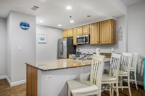 A kitchen with wooden cabinets, a stainless steel refrigerator, a microwave, and a granite countertop. There are white chairs and a small island with a sink. - 19th Floor Gulf Views | 2 King Suites | Splash Resort East Tower (Panama City Beach)
