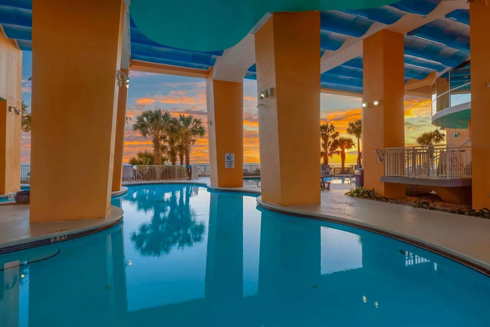 Piscina interior con vista al atardecer, rodeada de palmeras y una zona techada con asientos.