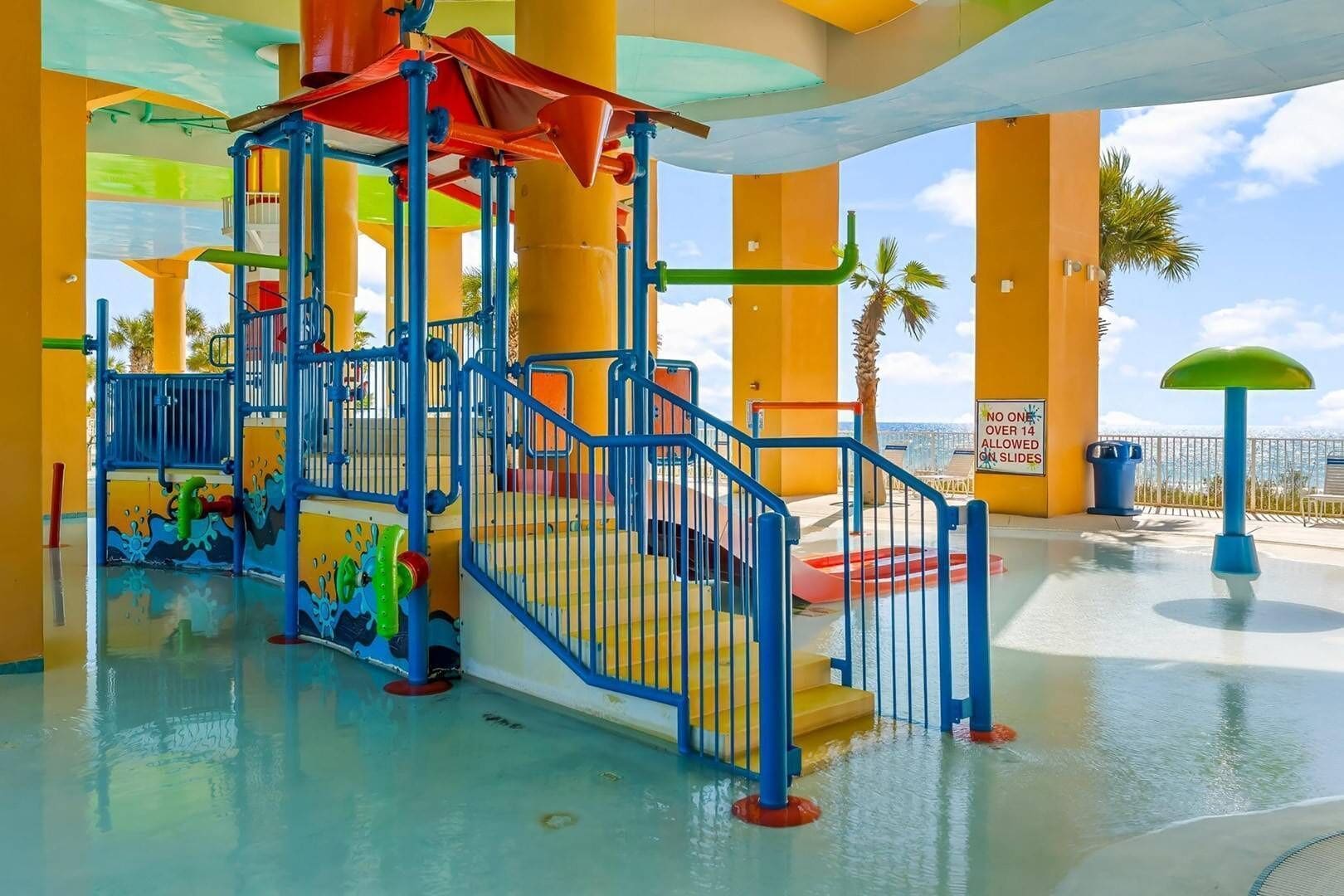 A water park with a colorful play structure, a no one over 14 allowed sign, and a view of palm trees and the sky.