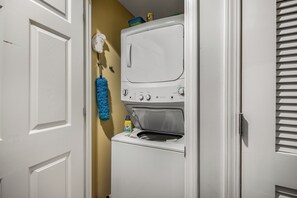 A compact laundry room with a front-loading washer and dryer, a blue mop, and a bottle of detergent. - 19th Floor Gulf Views | 2 King Suites | Splash Resort East Tower (Panama City Beach)