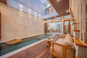 Een modern buitenzwembad met een stenen muur, een houten terras en een zithoek met een tafel en kussens.