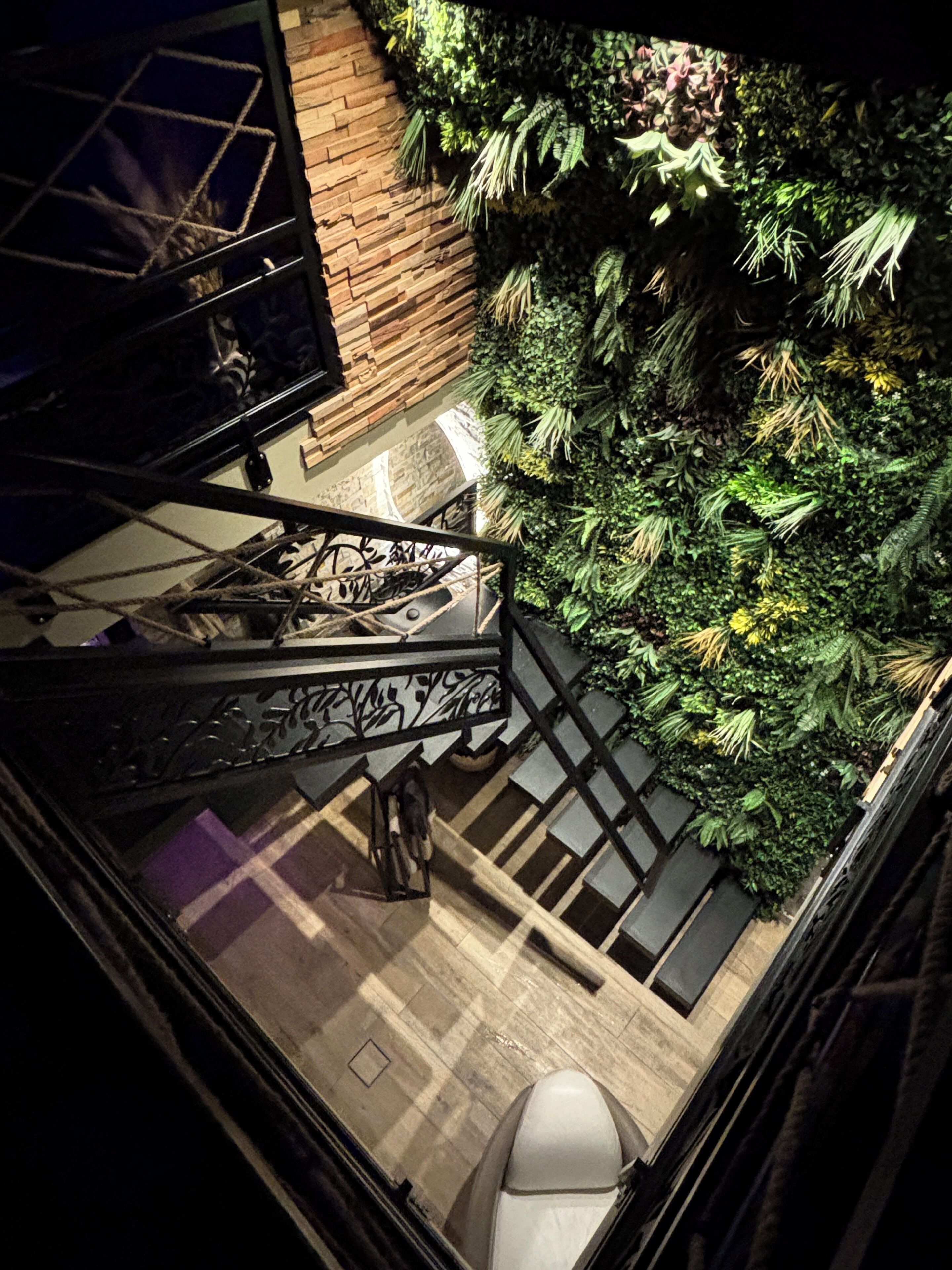 A staircase with a glass railing leading to a room with a wooden floor, a white chair, and a wall with green foliage.