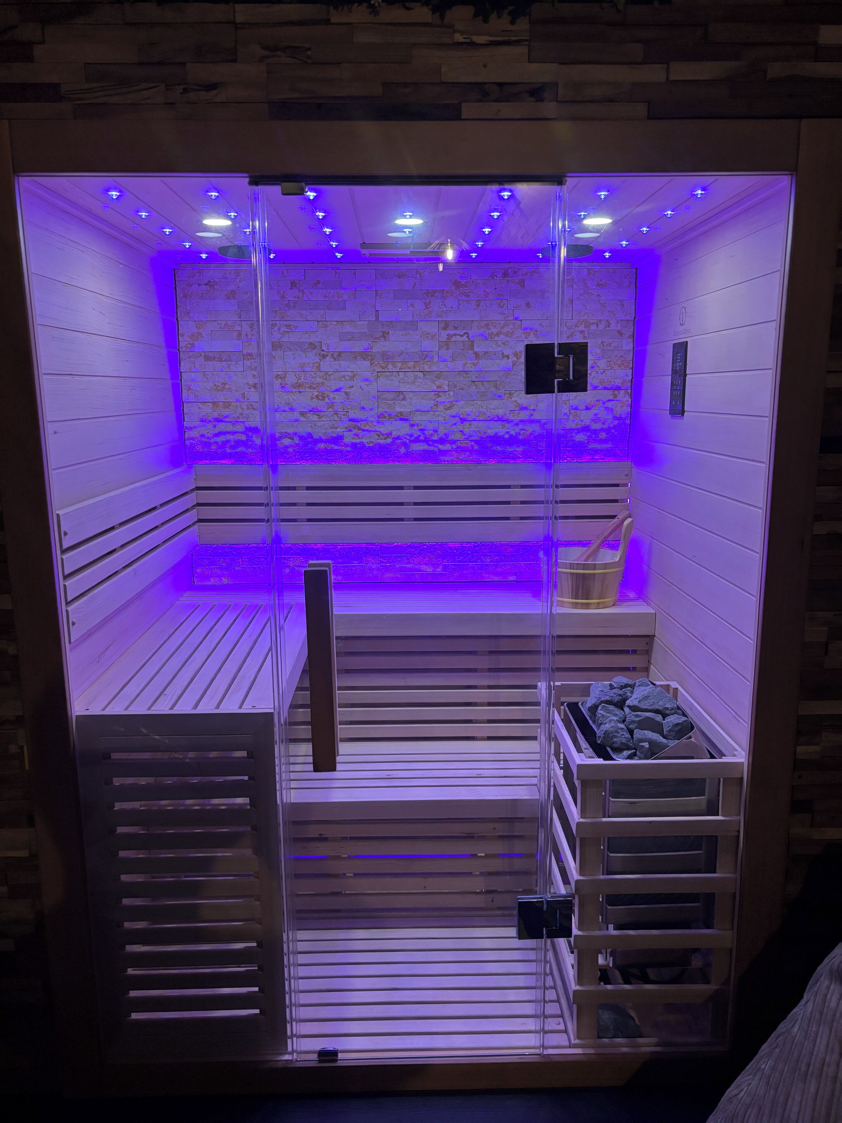 A sauna with wooden benches and a stone wall, illuminated by blue lighting.