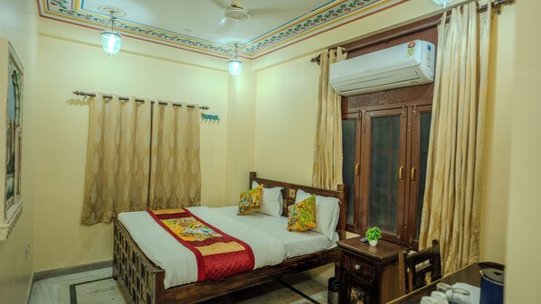 A bedroom with a bed, curtains, an air conditioning unit, a wooden wardrobe, and a small table with a plant. - BABA HAVELI HERITAGE HOTEL (Jaipur)