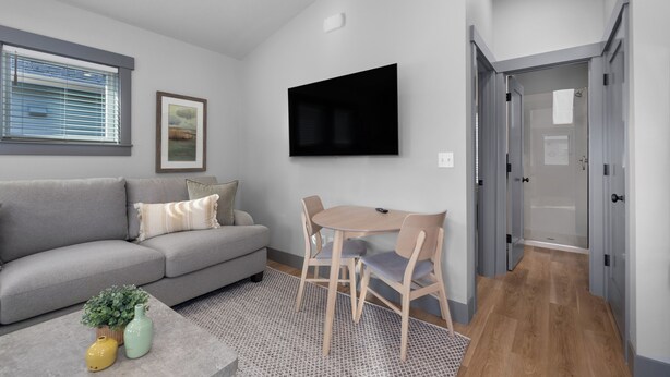 Ein modernes Wohnzimmer mit grauem Sofa, einem Couchtisch mit Dekoration, einem an der Wand montierten Fernseher und einer Glastür zu einem Duschbereich.