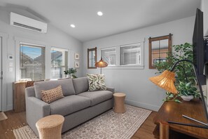 A modern living room with a grey sofa, a woven side table, a patterned rug, and a large window with blinds. - Seabrook Hazelwood Cottage | Pet Friendly, Kitchen | 1BR, Sleeps 2, Fernwood (Pacific Beach)