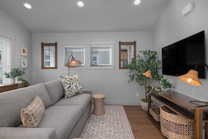 A living room with a grey sofa, two side tables, a flat-screen TV, and a large window with blinds. - Seabrook Hazelwood Cottage | Pet Friendly, Kitchen | 1BR, Sleeps 2, Fernwood (Pacific Beach)