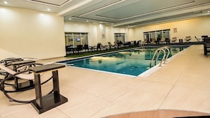 Una piscina con sillas de descanso y mesas, piso de baldosas y una pantalla de televisión.