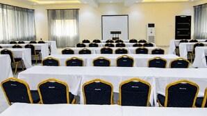 Un salón de conferencias con filas de mesas y sillas, una pantalla de proyector y una pizarra.
