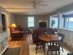 A living room with a red sofa, a wooden dining table with chairs, a ceiling fan, and a television.