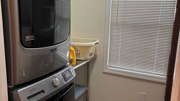 A compact kitchen with a stainless steel refrigerator, a built-in oven, and a microwave. There is a window with blinds, a laundry basket, and a yellow bottle on a shelf.