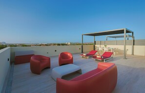 Eine Dachterrasse mit roten und weißen Liegestühlen, einer überdachten Sitzgelegenheit und Blick auf die Stadt.