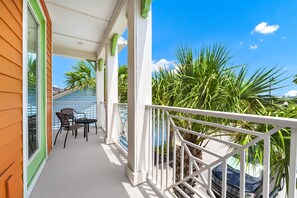 A balcony with a white railing, a table, two chairs, and a view of palm trees and a building. - Tropical Cottage near Disney | Resort & Waterpark Access - 3095KL (Kissimmee)