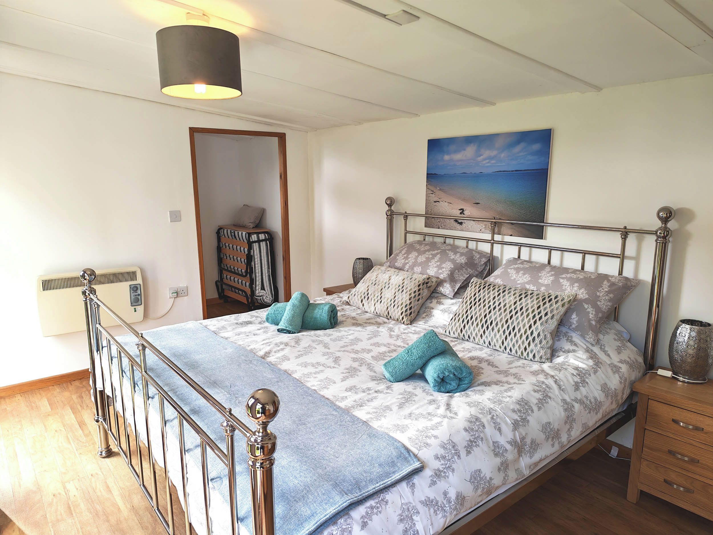 A bedroom with a metal-framed bed, a wall-mounted heater, a painting of a beach, and a wooden nightstand.
