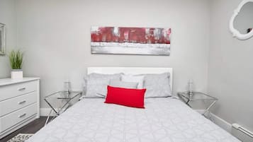 Een moderne slaapkamer met een wit bed, grijs en rood kussen, een rood accentkussen, en een groot abstract schilderij aan de muur.
