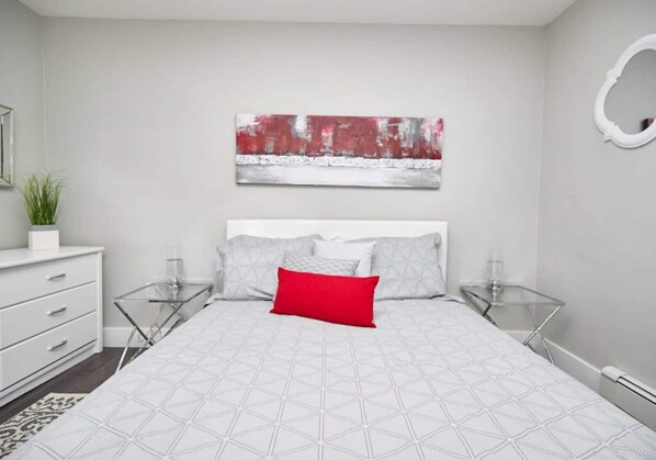 A modern bedroom with a white bed, grey and red pillows, a red accent pillow, and a large abstract painting on the wall.