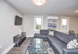 A modern living room with a grey sectional sofa, a glass coffee table, and a flat-screen TV mounted on the wall. There are two windows with air conditioning units above them, and a painting is visible on the wall.
