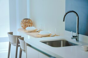Une cuisine moderne avec un plan de travail blanc, un évier en acier inoxydable et une étagère en bois où sont posées des assiettes et un verre.