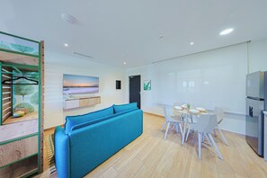 A modern living room with a blue sofa, a dining table with chairs, a kitchen area with a refrigerator, and a wall-mounted TV displaying a sunset scene. - Bonsai Studio-Leaf 203 (Noord)