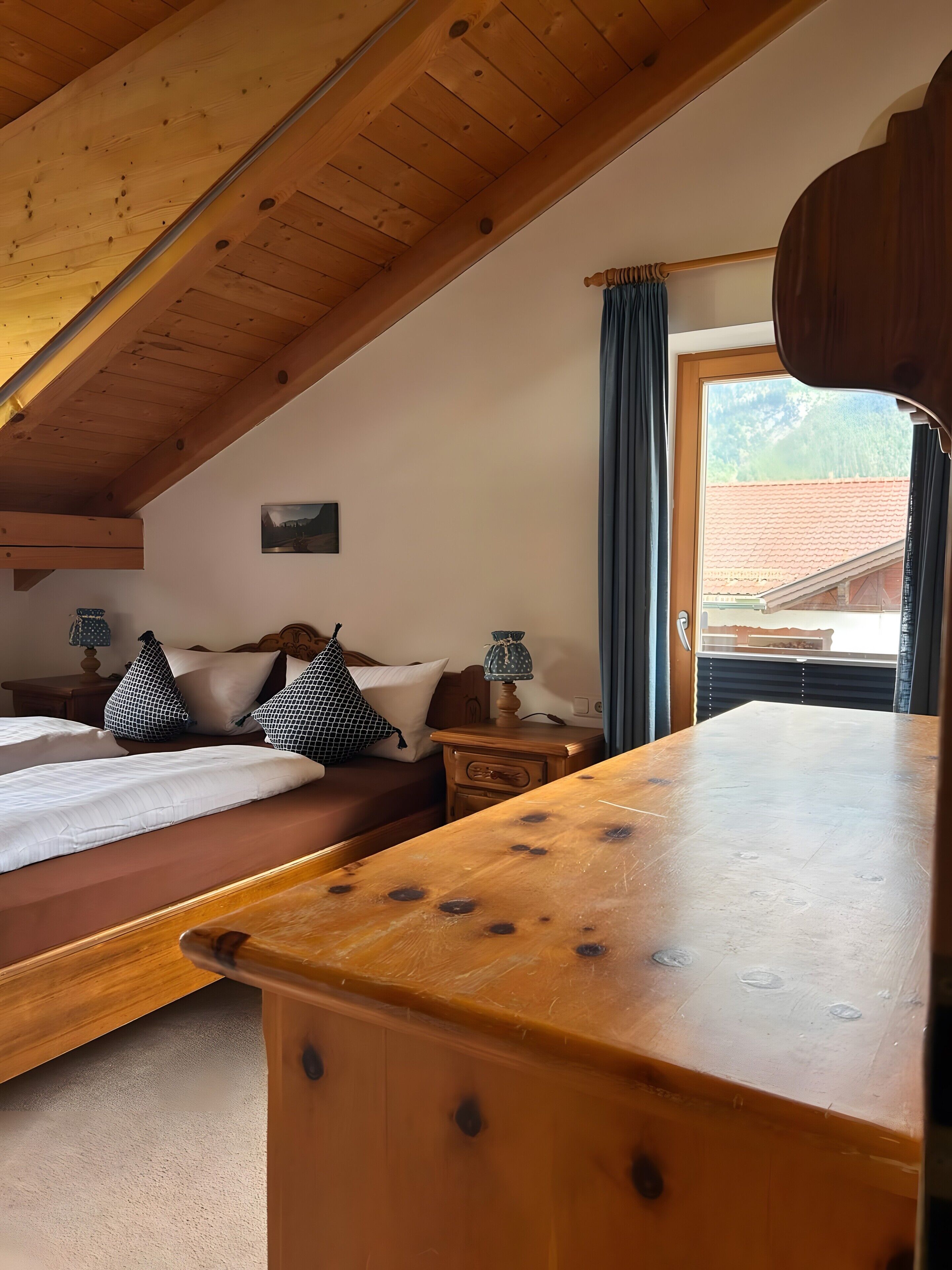 Una habitación acogedora con techo de madera, cama con almohadas blancas y estampadas, mesita de noche con lámpara y vista a la montaña a través de una ventana.