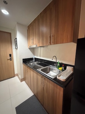 A kitchen with wooden cabinets, a black countertop, stainless steel sink, and a black refrigerator. - Prime MOA Complex 1 BR Corner Condo Unit - Walk to Mall, Restaurants & the Bay (Pasay City)