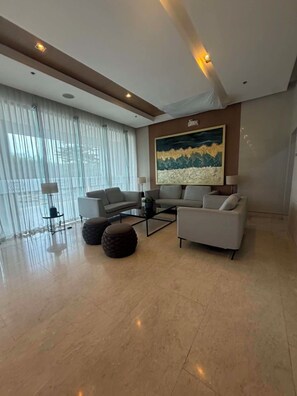 A modern living room with a sofa, coffee table, and ottomans, large windows with sheer curtains, and a painting on the wall. - Prime MOA Complex 1 BR Corner Condo Unit - Walk to Mall, Restaurants & the Bay (Pasay City)
