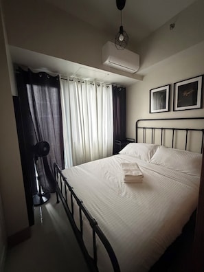 A hotel room with a bed, a standing fan, an air conditioning unit, a window with curtains, and two framed pictures on the wall. - Prime MOA Complex 1 BR Corner Condo Unit - Walk to Mall, Restaurants & the Bay (Pasay City)
