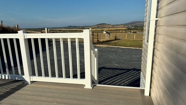 A wooden deck with a white railing, a clear view of a fenced pasture with cows, and a distant landscape. - Bwlchgwyn Caravan, Aberdaron (Pwllheli)