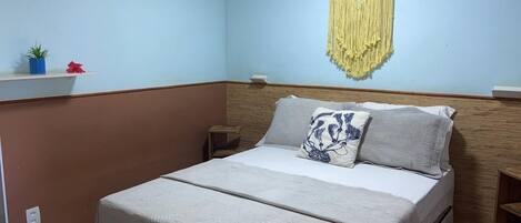 A single bed with a patterned bedspread, a bedside table, a wall-mounted macrame decoration, and a small shelf with a potted plant and a red flower.