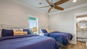 Ein Schlafzimmer mit zwei Betten, einem Deckenventilator, einem Fenster mit Jalousien sowie einem Badezimmer mit Spiegel und Waschbecken.