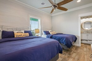 Ein Schlafzimmer mit zwei Betten, einem Deckenventilator, einem Fenster mit Jalousien sowie einem Badezimmer mit Spiegel und Waschbecken.