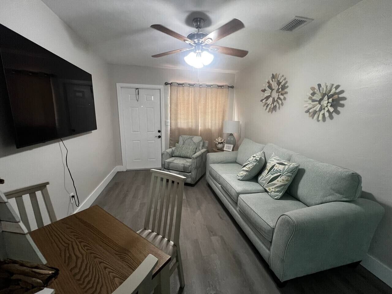 A living room with a ceiling fan, a television mounted on the wall, a sofa, a chair, and a dining table.